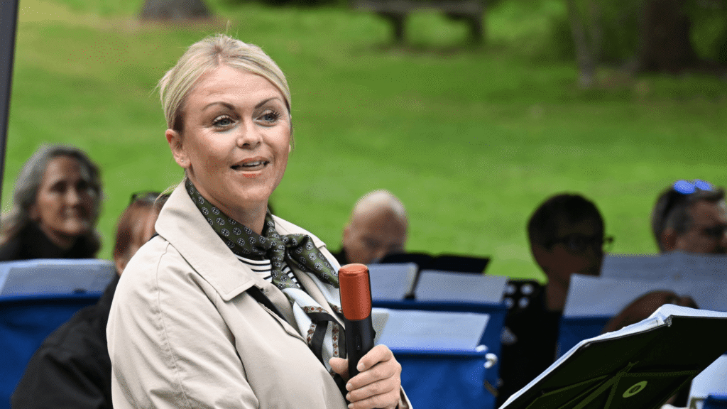 A lady in a cream coat holds a microphone while talking to a crowd of people.