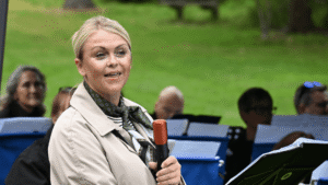 A lady in a cream coat holds a microphone while talking to a crowd of people.