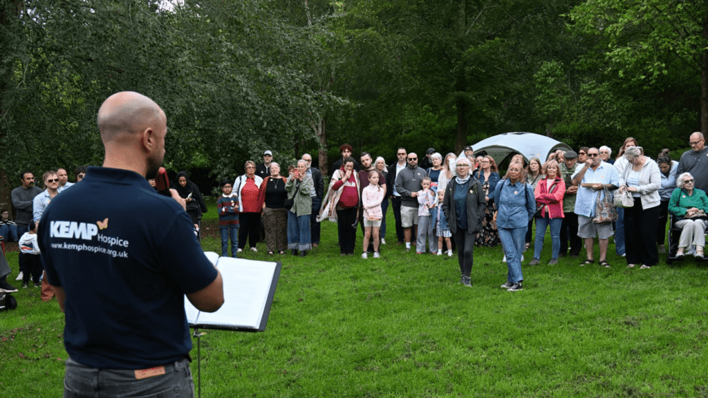 a man stands with his back to the camera while holding a microphone while speaking to a crowd of people