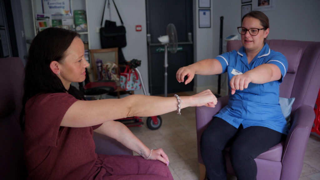 a nurse in blue scrubs sits opposite a lady in a purple tip with arms outstretched showing an exercise