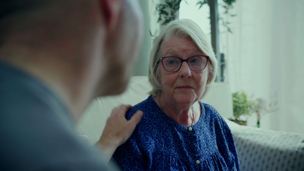 a person sits looking towards another person whose hand is on her left shoulder