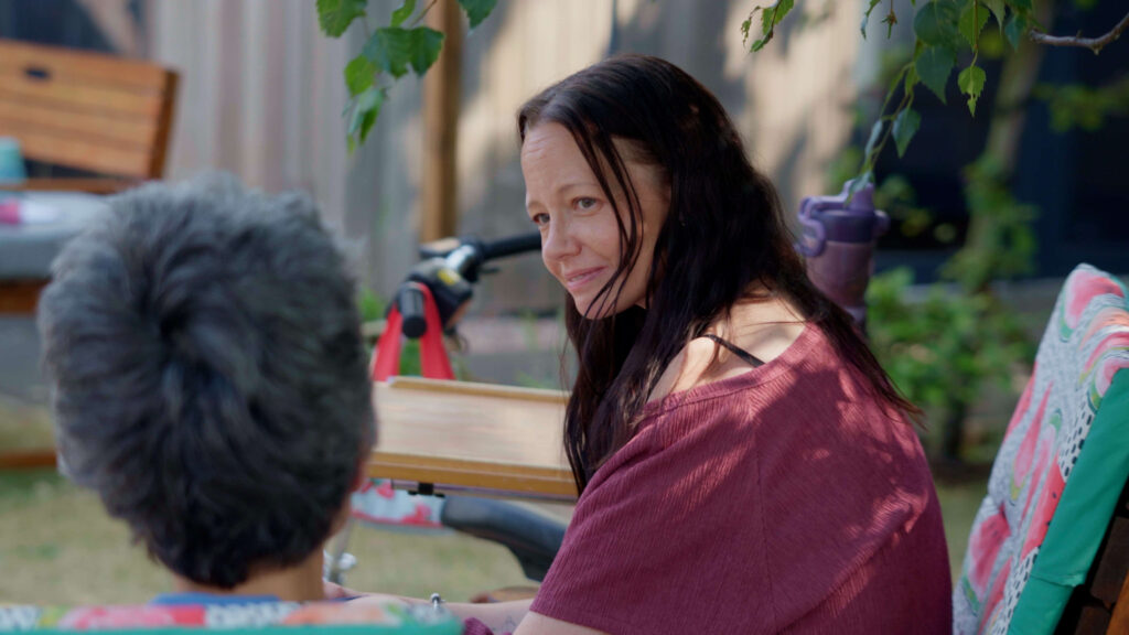 a lady with a purple top sits in a garden talking to another person sat on a chair